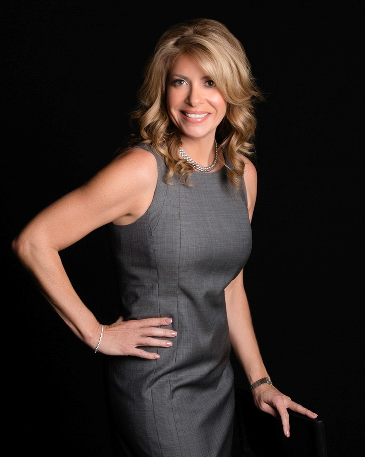 Woman with blonde hair in a gray sleeveless dress, hand on hip, dark backdrop, photographed by Annie Watson in her Naples studio