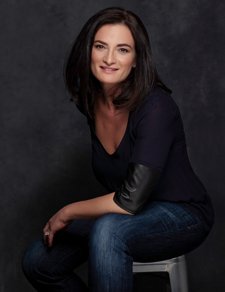 Woman with dark hair in a black top and jeans, seated on a stool, dark backdrop, photographed by Annie Watson in her Naples studio